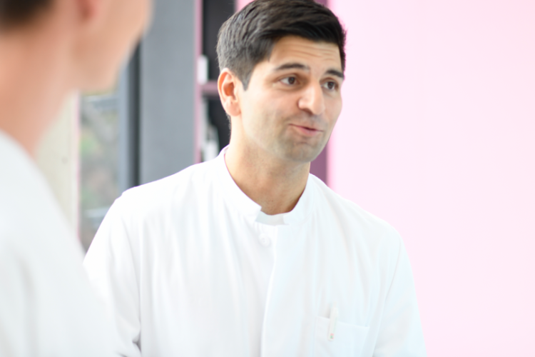 Ein männlicher Mitarbeiter in weißer medizinischer Berufskleidung spricht mit Kolleg*innen in einem hellen Raum.