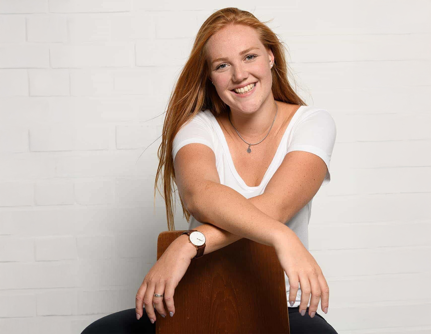 Junge Frau mit roten Haaren sitzt entspannt rückwärts auf einem Stuhl und lächelt in die Kamera.