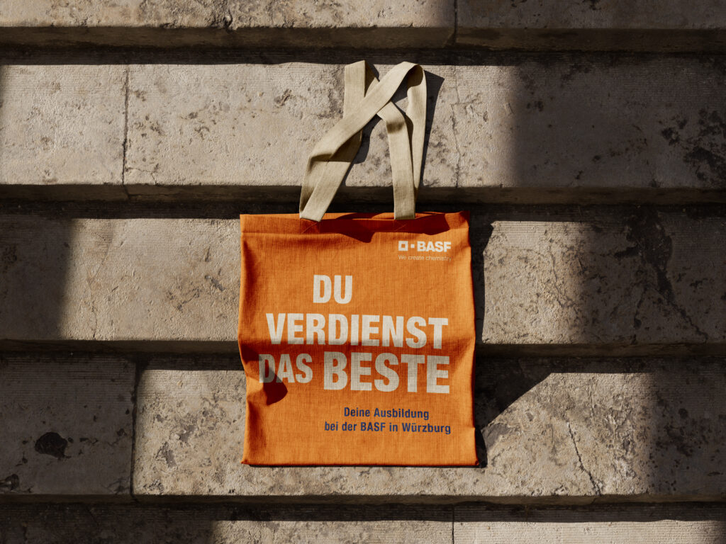 Orange Stofftasche mit dem Claim „Du verdienst das Beste“ aus der BASF-Azubi-Recruiting-Kampagne – fotografiert als Employer-Branding-Merchandising im urbanen Umfeld.