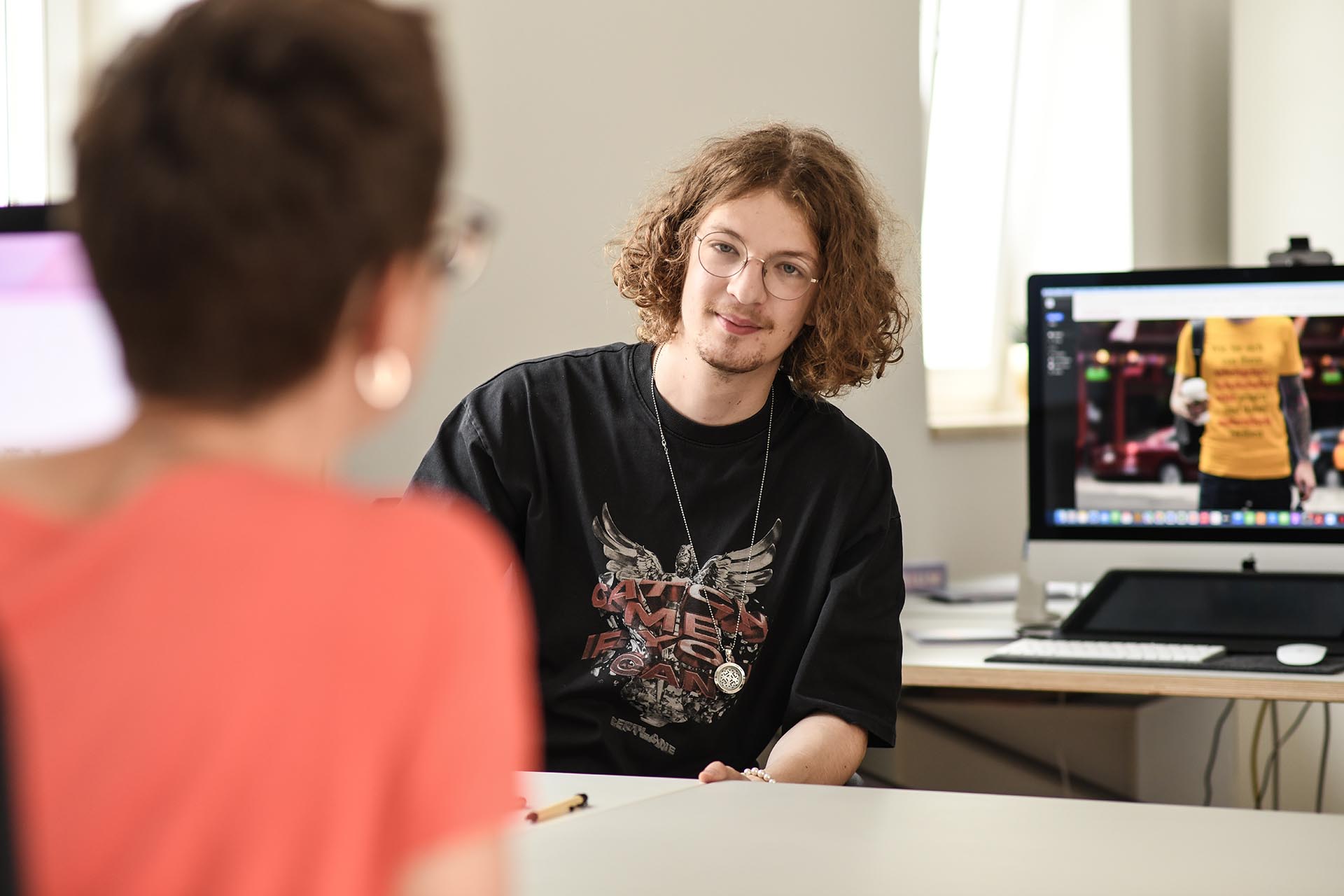 Junger Mitarbeiter der Agentur Heldenstreich im Gespräch mit einer Kollegin im Büro, vor einem Bildschirm mit Design-Layout – authentischer Einblick in die kreative Zusammenarbeit.