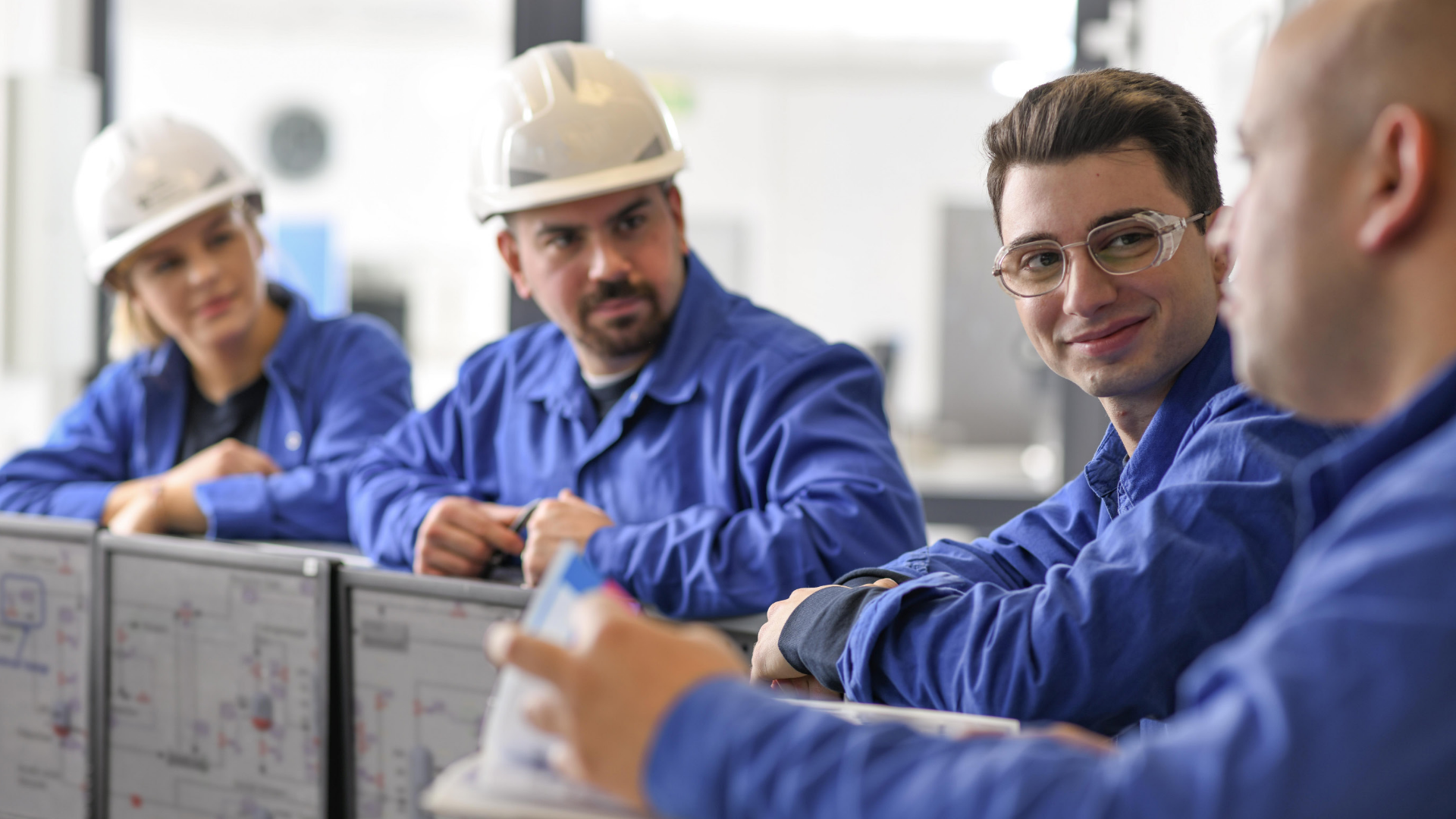 Mitarbeiter von Boehringer Ingelheim in blauer Arbeitskleidung im Gespräch an Monitoren in einem technischen Arbeitsumfeld.