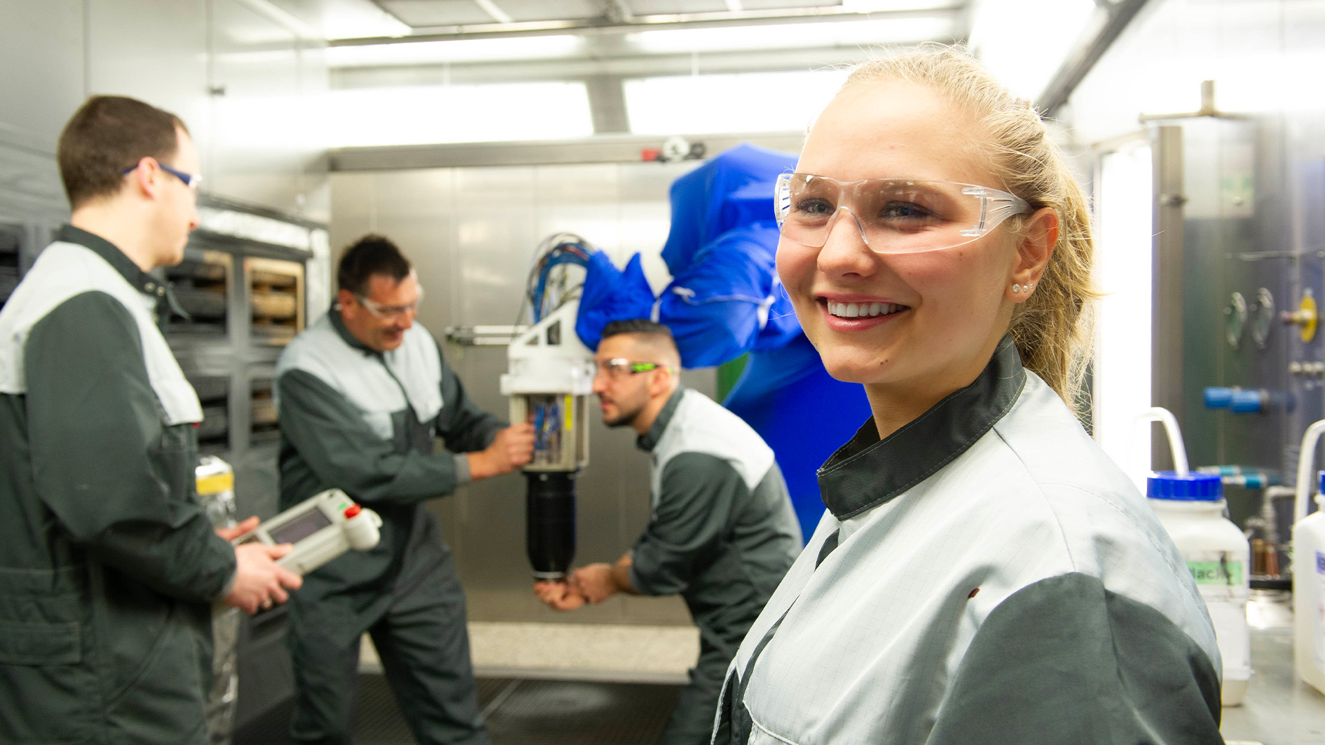 Azubis der BASF Coatings Würzburg arbeiten gemeinsam in der Lackierkabine; eine junge Mitarbeiterin steht lächelnd im Vordergrund.