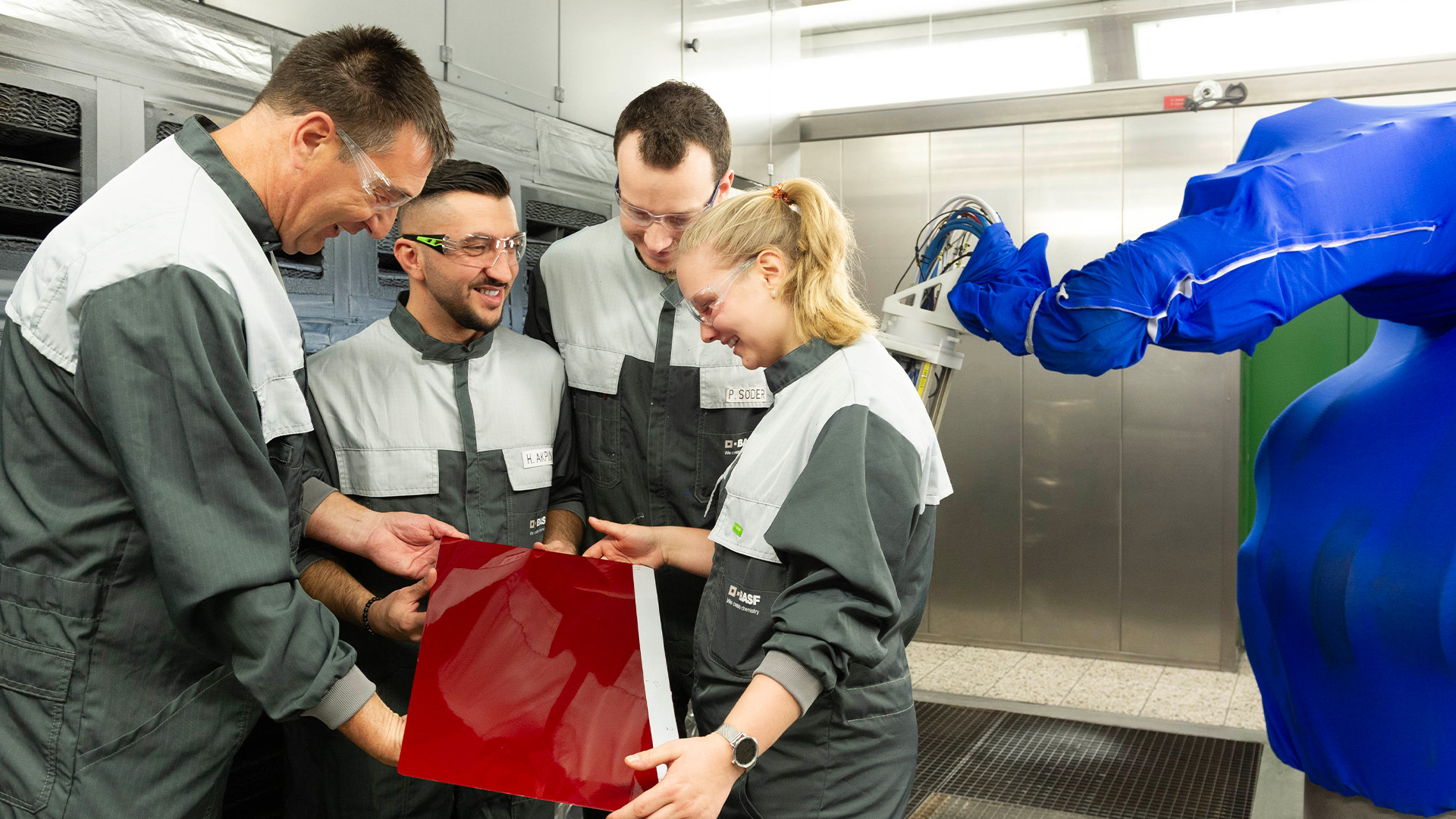 Gruppe von Mitarbeitenden in Schutzkleidung betrachtet gemeinsam eine lackierte rote Platte in der Produktion bei BASF Coatings Würzburg.
