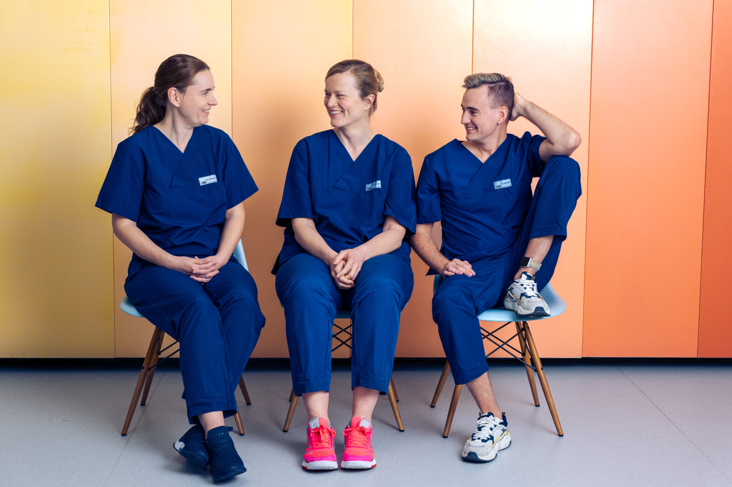 Drei Pflegekräfte des LMU Klinikums sitzen gemeinsam vor einer orange-gelben Wand und lächeln in die Kamera.