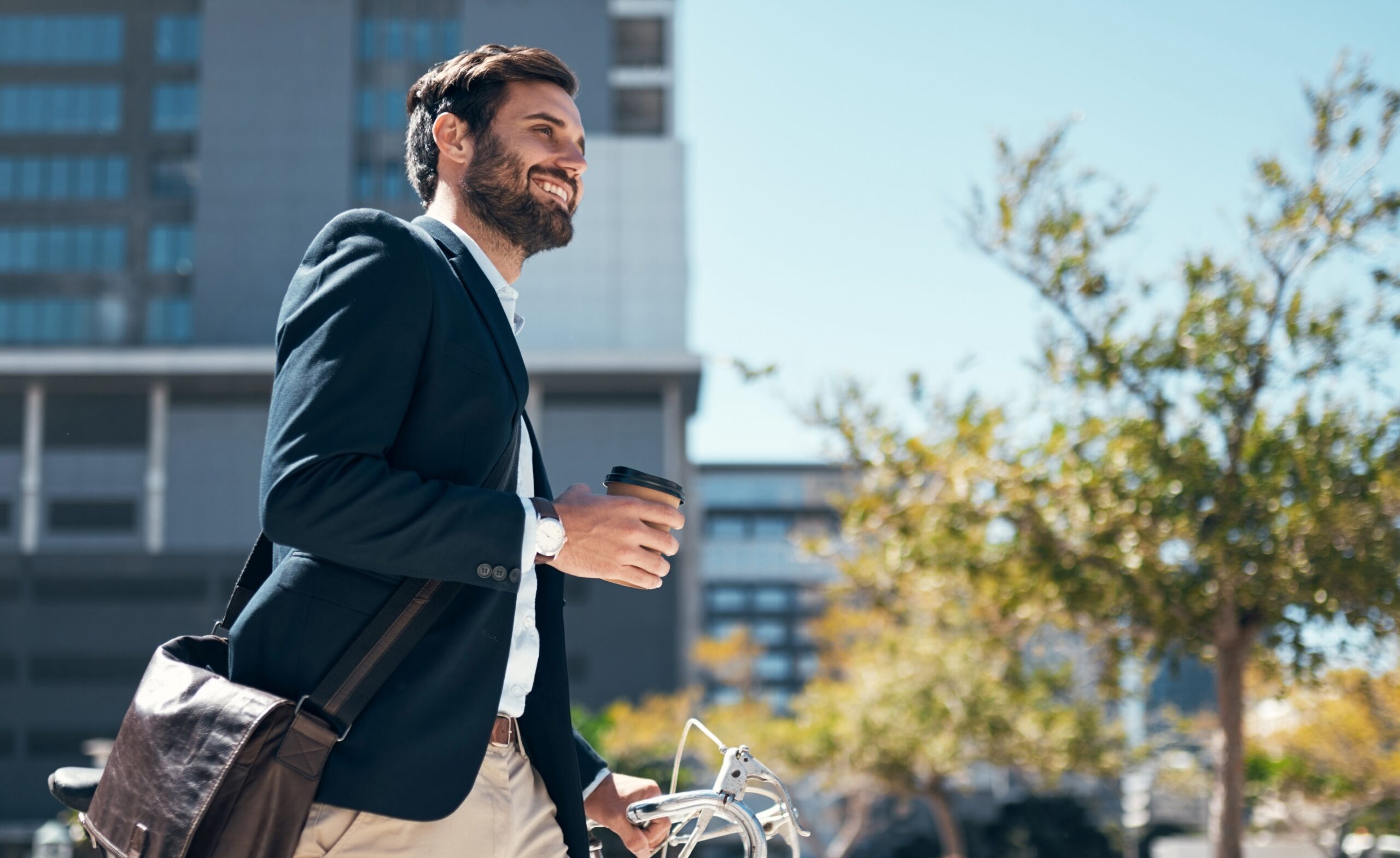 Mann im Business-Outfit mit Kaffeebecher und Fahrrad in moderner Stadtumgebung – Keyvisual der Kampagne zum MBA Internationales Immobilienmanagement.