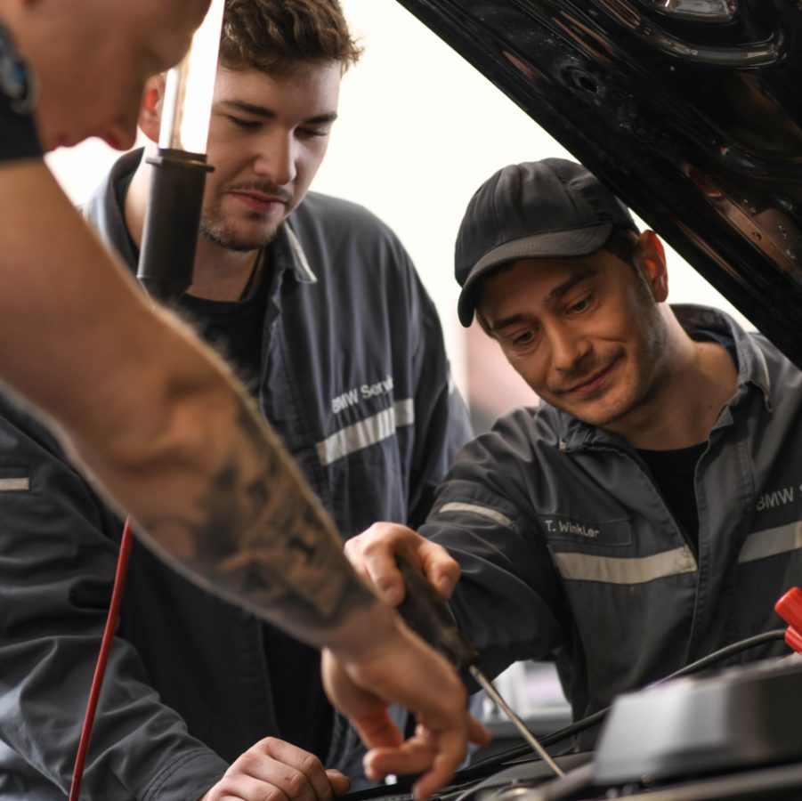 Mechaniker:innen des BMW Service Teams arbeiten gemeinsam unter der Motorhaube und überprüfen ein Fahrzeug.