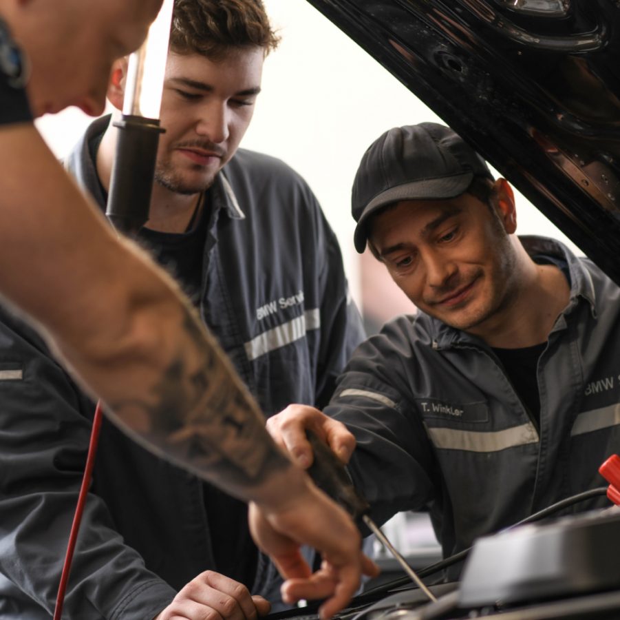 Mechaniker:innen des BMW Service Teams arbeiten gemeinsam unter der Motorhaube und überprüfen ein Fahrzeug.