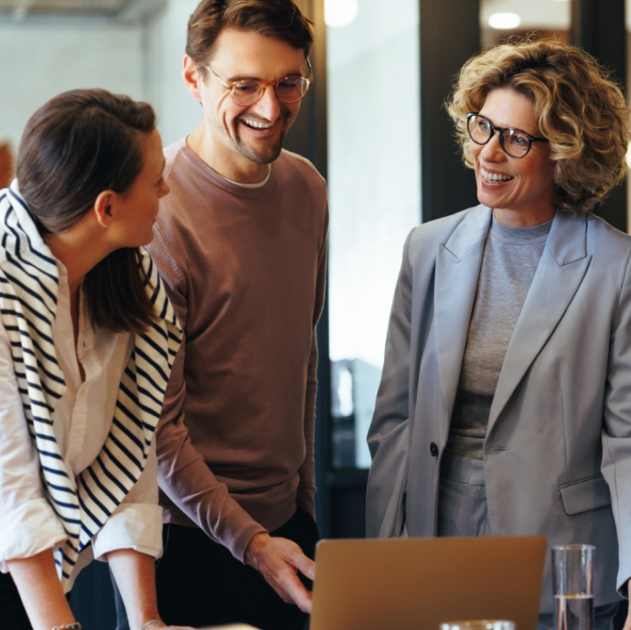 „Zwei studierende Personen und eine Professorin stehen gemeinsam in einem modernen Büro, lachen und arbeiten an einem Laptop – Symbolbild für Teamwork und berufliche Weiterbildung.“