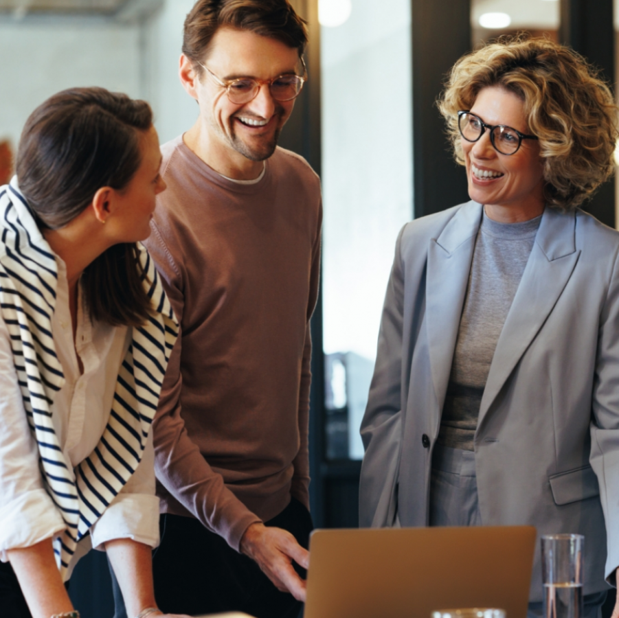 „Zwei studierende Personen und eine Professorin stehen gemeinsam in einem modernen Büro, lachen und arbeiten an einem Laptop – Symbolbild für Teamwork und berufliche Weiterbildung.“