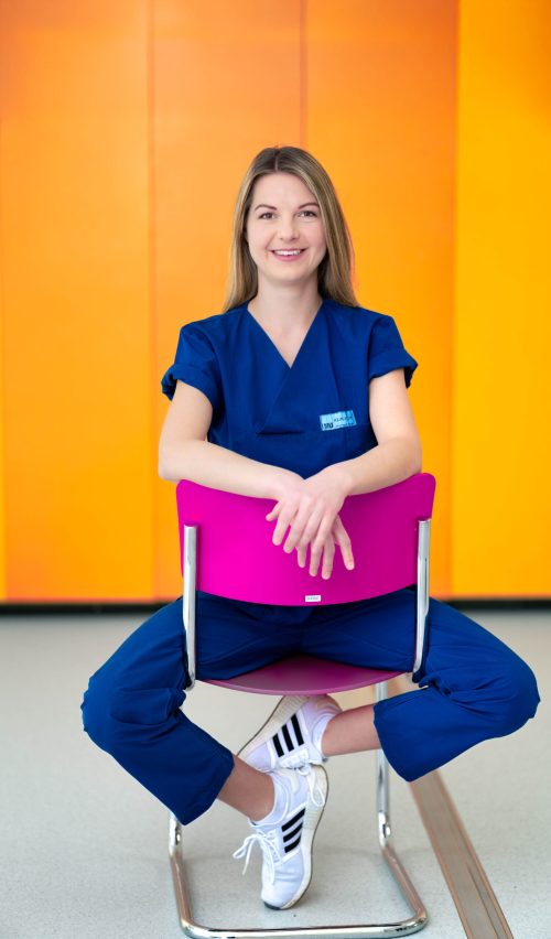 Pflegefachkraft des LMU Klinikums sitzt entspannt auf einem pinken Stuhl vor einer orangefarbenen Wand und lächelt freundlich in die Kamera.