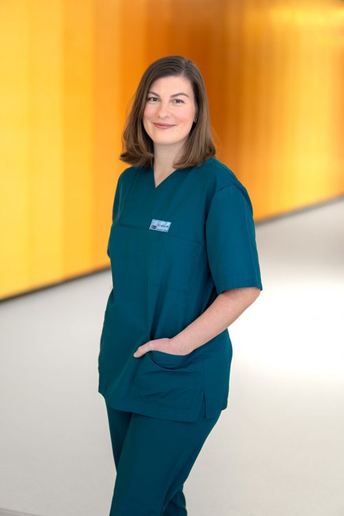 Pflegekraft in türkiser Arbeitskleidung steht lächelnd vor einem warmen, gelb-orangen Hintergrund im Klinikum.