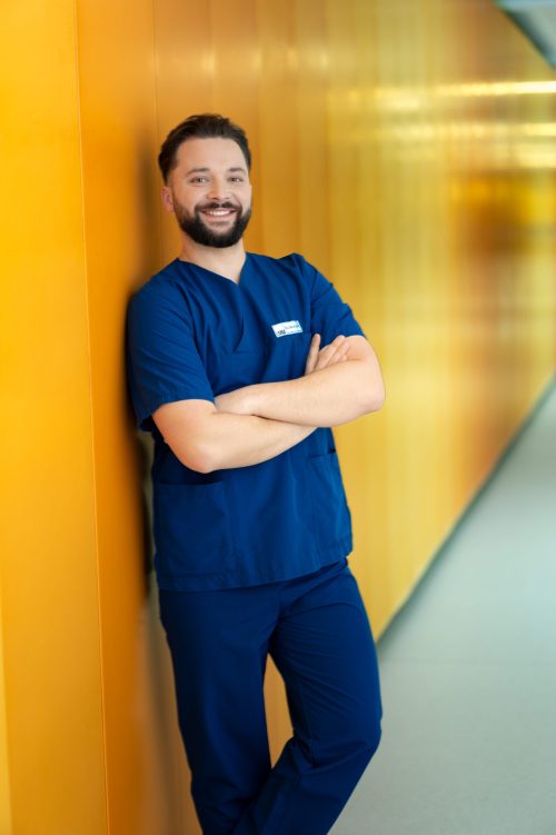 Männlicher Mitarbeiter des LMU Klinikums in blauer Pflegekleidung lehnt lächelnd an einer gelben Wand