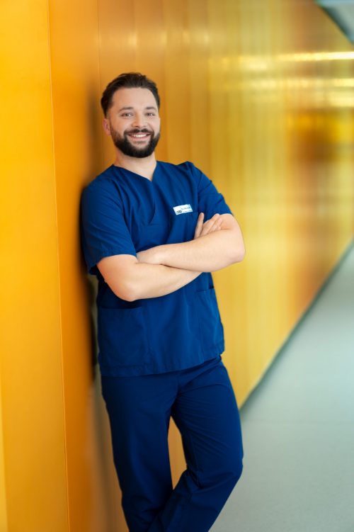 Männlicher Mitarbeiter des LMU Klinikums in blauer Pflegekleidung lehnt lächelnd an einer gelben Wand