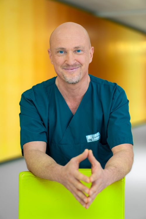 Pflegefachkraft des LMU Klinikums lächelnd im Porträt vor warmem gelbem Hintergrund.