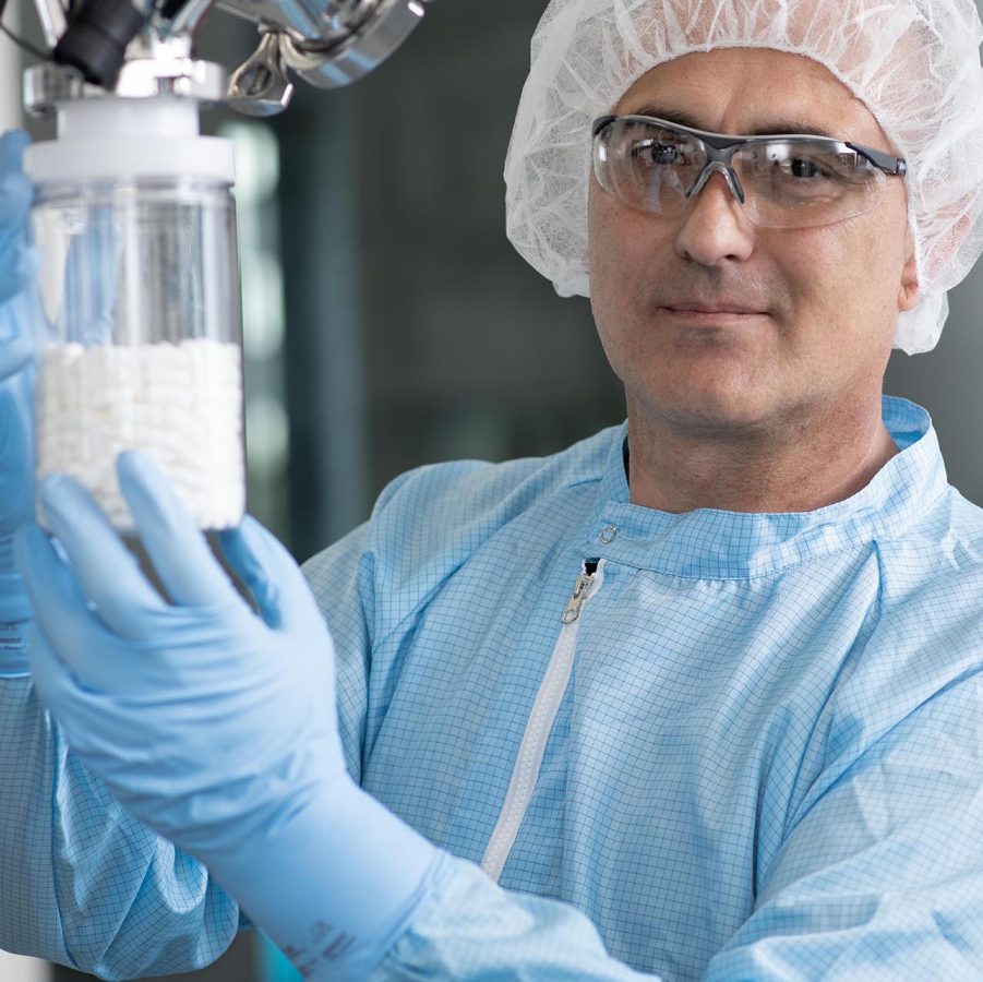 Mitarbeiter in Schutzkleidung hält ein Glas mit weißen Tabletten in einer pharmazeutischen Produktionsanlage.